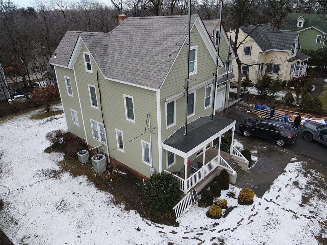 Aerial view of a green two-story house with a porch, parked cars, and snowy surroundings.